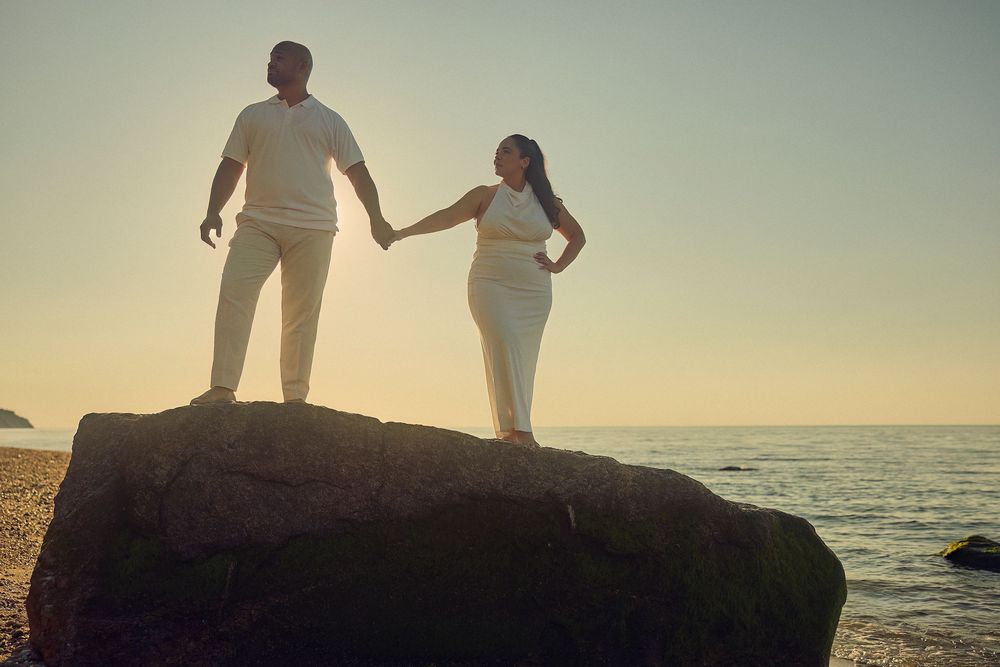 Two people hold hands while standing on coastal rocks at sunset with ocean waves crashing in the background.
