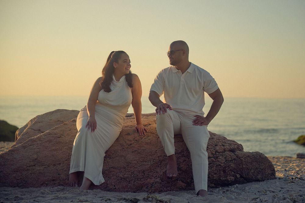 A couple shares a romantic moment at sunset on a rocky beach overlook with ocean waves in the background.