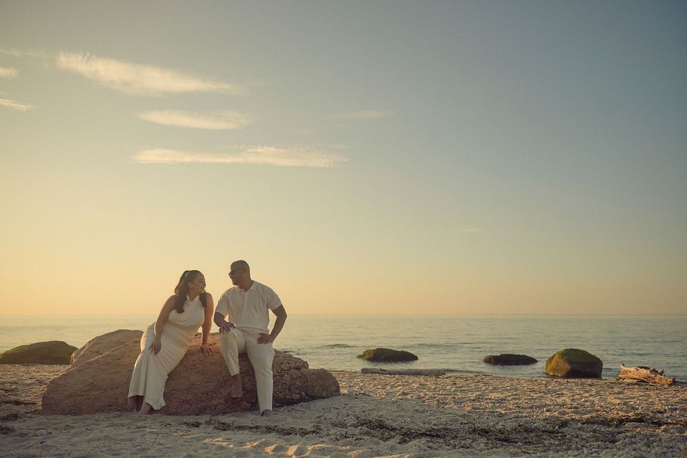 A couple sits together on coastal rocks at sunset overlooking a peaceful ocean horizon.