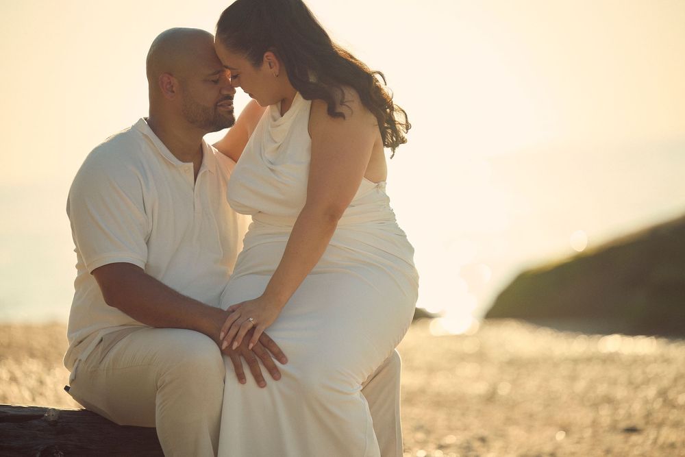 A romantic couple in white outfits sharing an intimate moment on the beach at sunset.