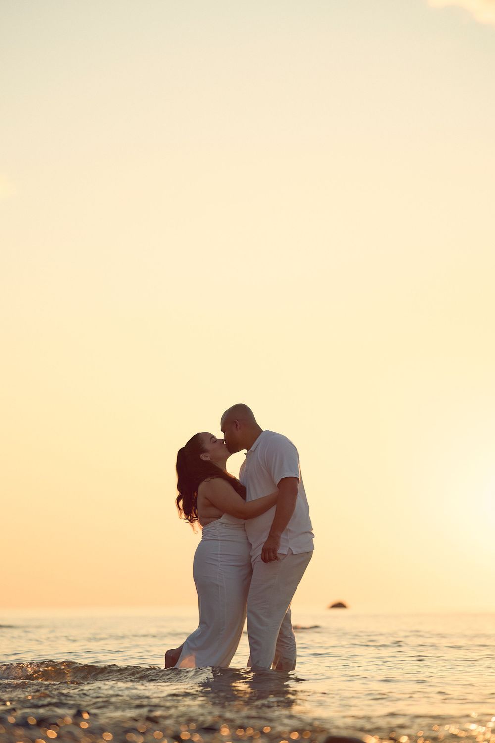 Romantic couple in white clothes embrace on beach at sunset by the ocean shore in tropical paradise.