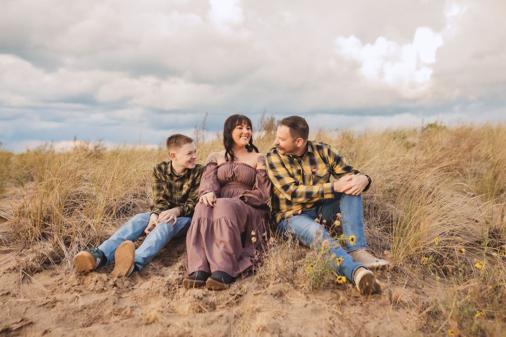 Family of three sits together on golden grassy hillside under cloudy sky during outdoor photo session.