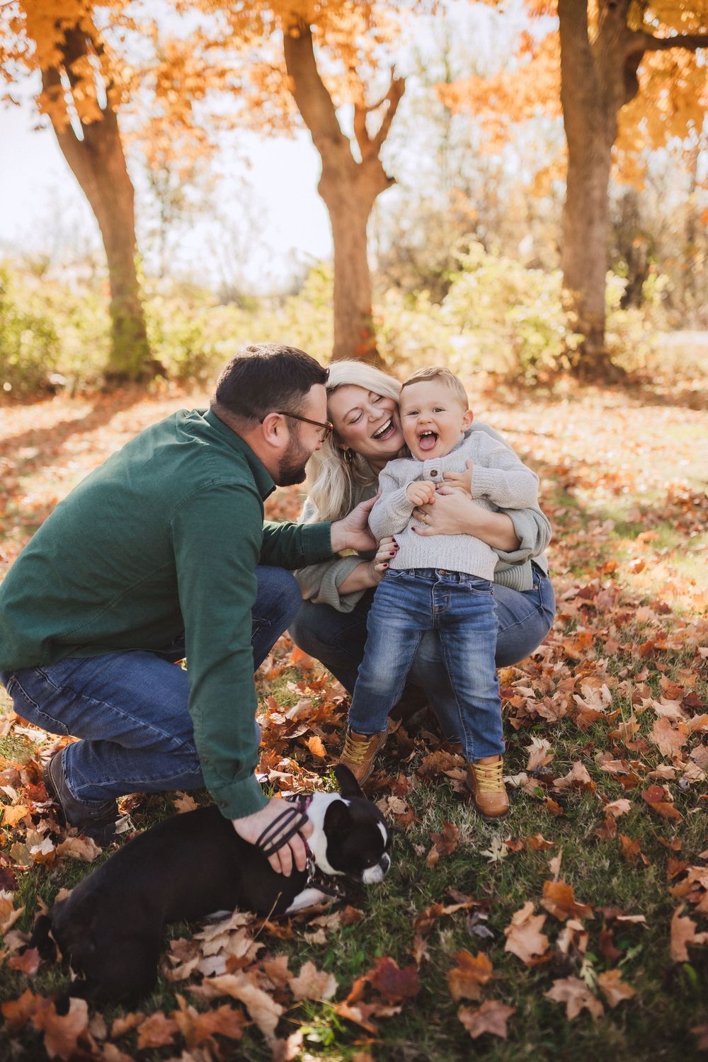 A family and their dog share a playful autumn moment among fallen orange leaves in a sunlit forest setting.