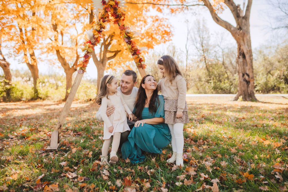 A group embraces while sitting on fallen autumn leaves beneath vibrant orange maple trees in a sunlit park.