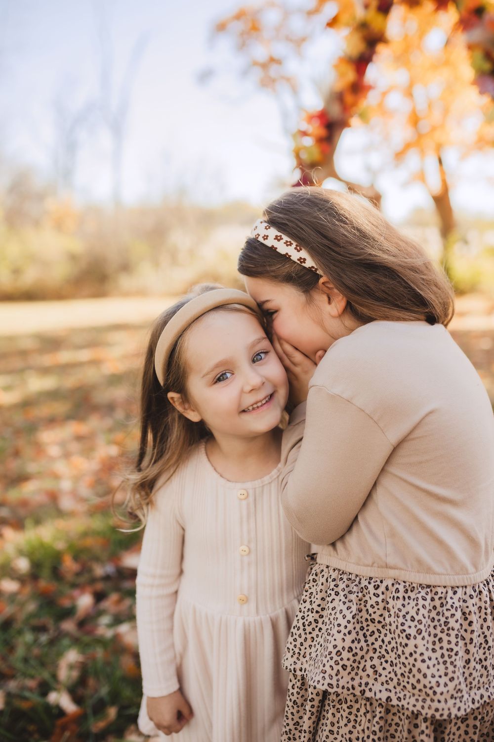 Two children in cream-colored dresses share a tender moment among autumn leaves in a dreamy outdoor setting.