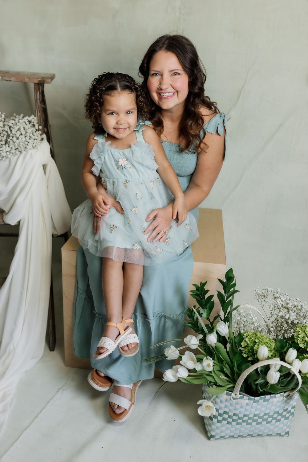 Two people in matching light blue dresses pose together against a neutral backdrop with white flowers and decor.