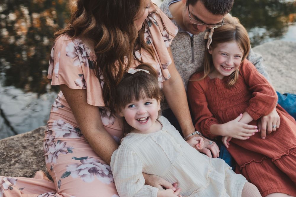 Family shares laughter during a casual outdoor photo session by the water in floral and solid colored dresses.