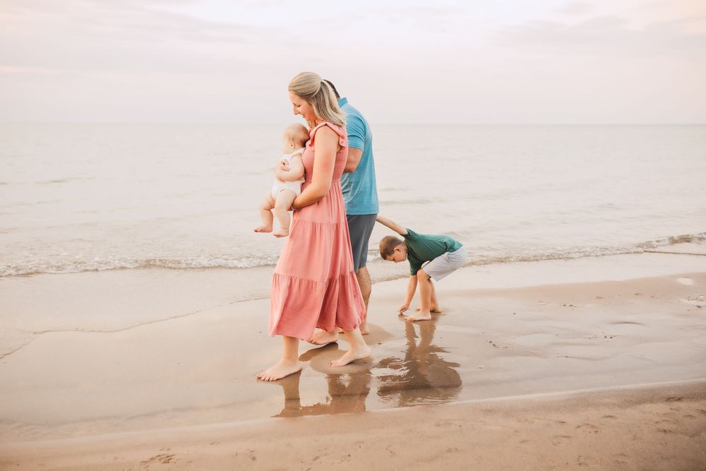 A parent and two children play in the sand at the beach during sunset, wearing colorful summer clothing.