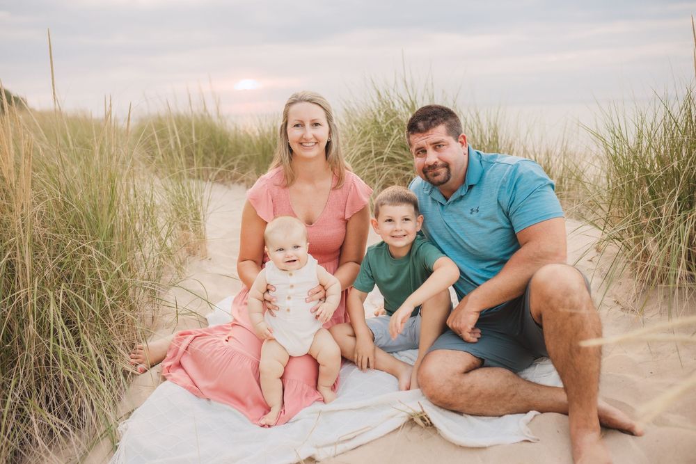 A family sits together on a blanket at the beach among sand dunes during sunset.