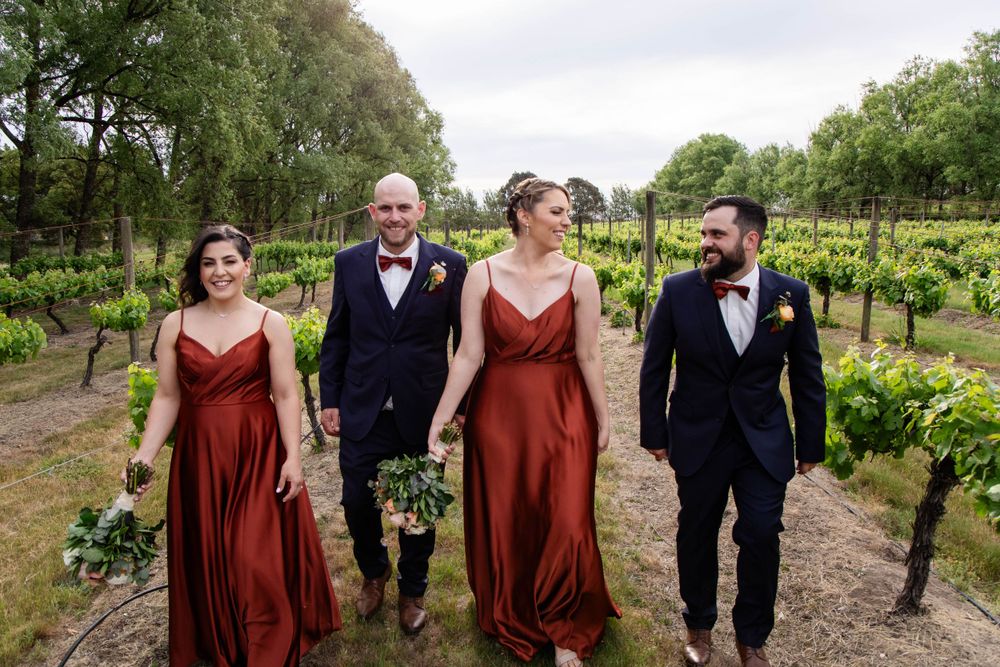 Wedding party walks through vineyard wearing burgundy dresses and navy suits.