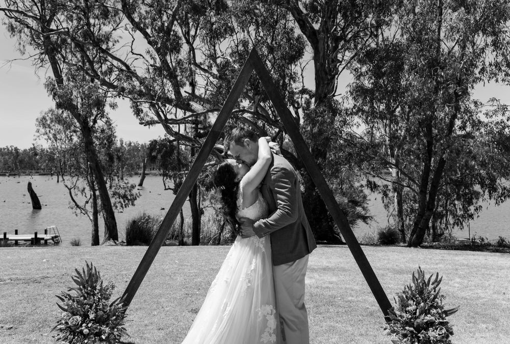Black and white photo of couple embracing under wooden triangle arch.