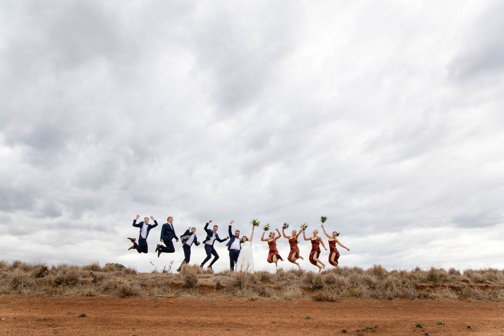 Wedding party jumps in celebration against dramatic cloudy sky in outback setting.