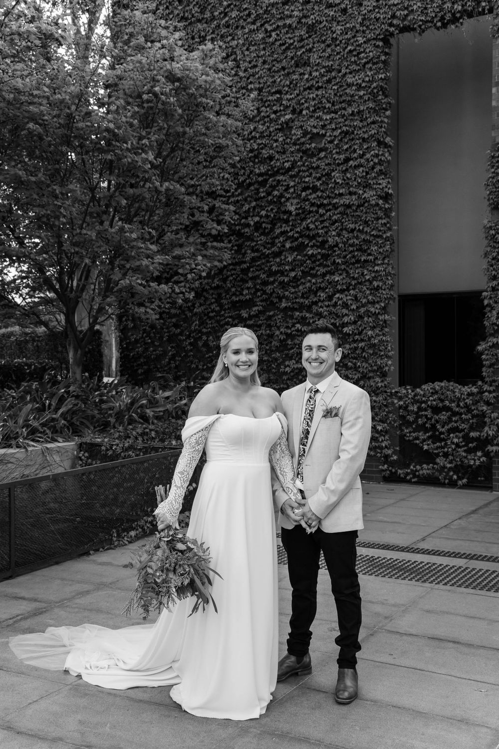 A wedding couple poses against an ivy-covered wall backdrop with a natural garden setting.