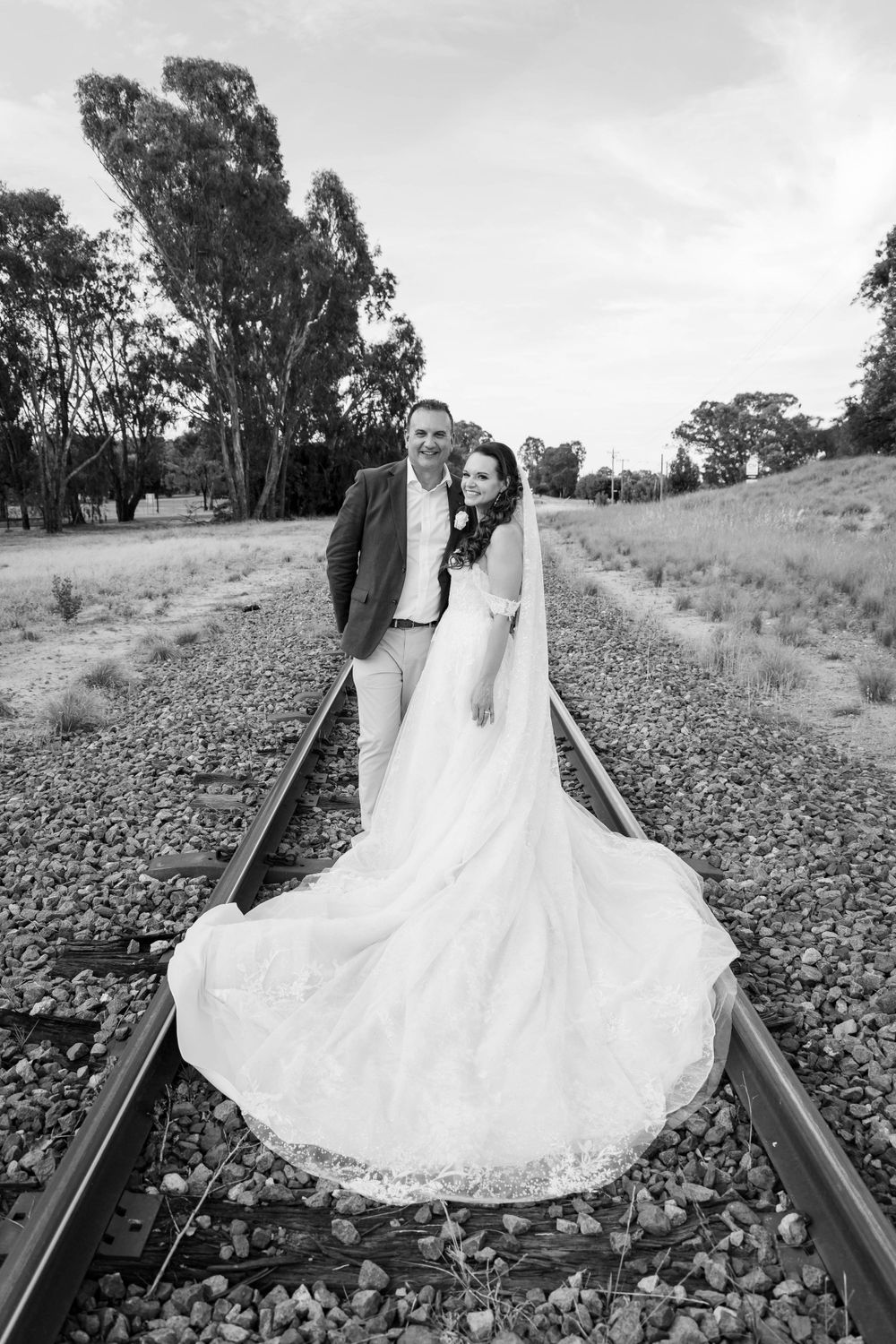 A wedding couple poses on train tracks with a flowing dress trail in black and white.
