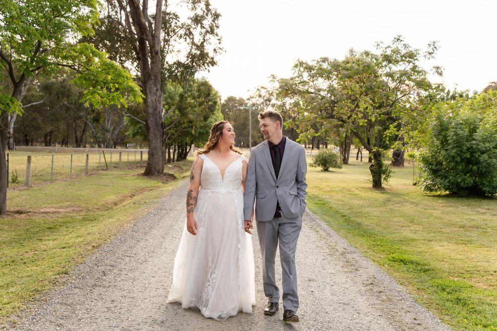 Couple walks hand in hand down a tree-lined gravel path on their wedding day.