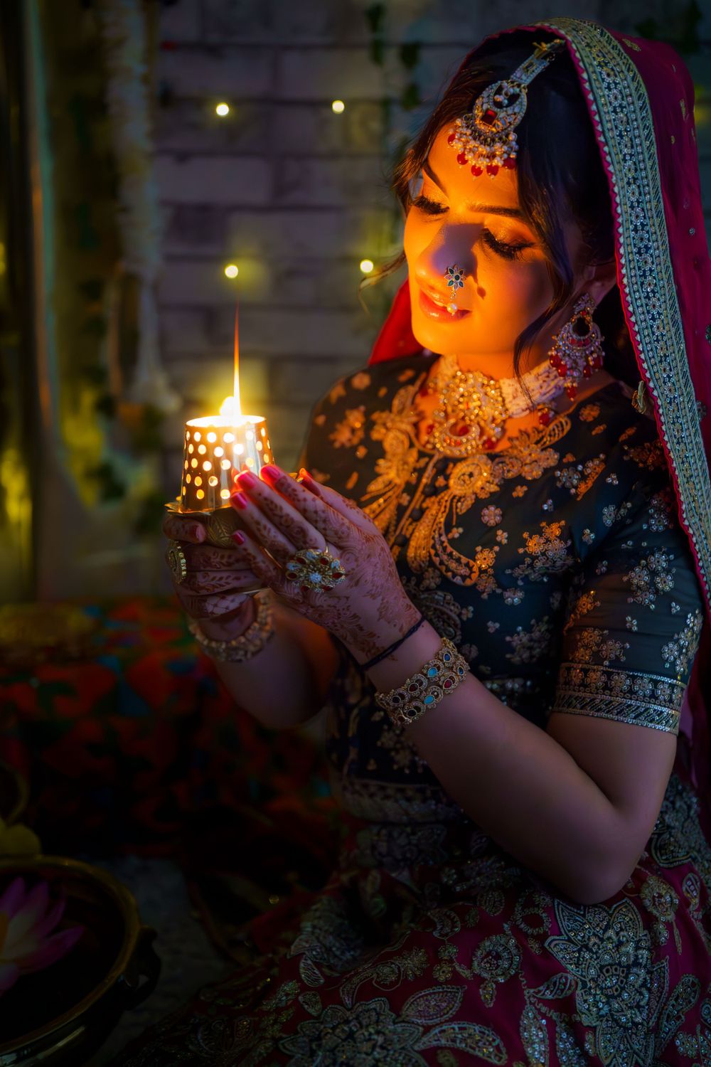 Traditional Indian bride in ornate dress and jewelry holds glowing candle in evening, with bokeh lights in background.