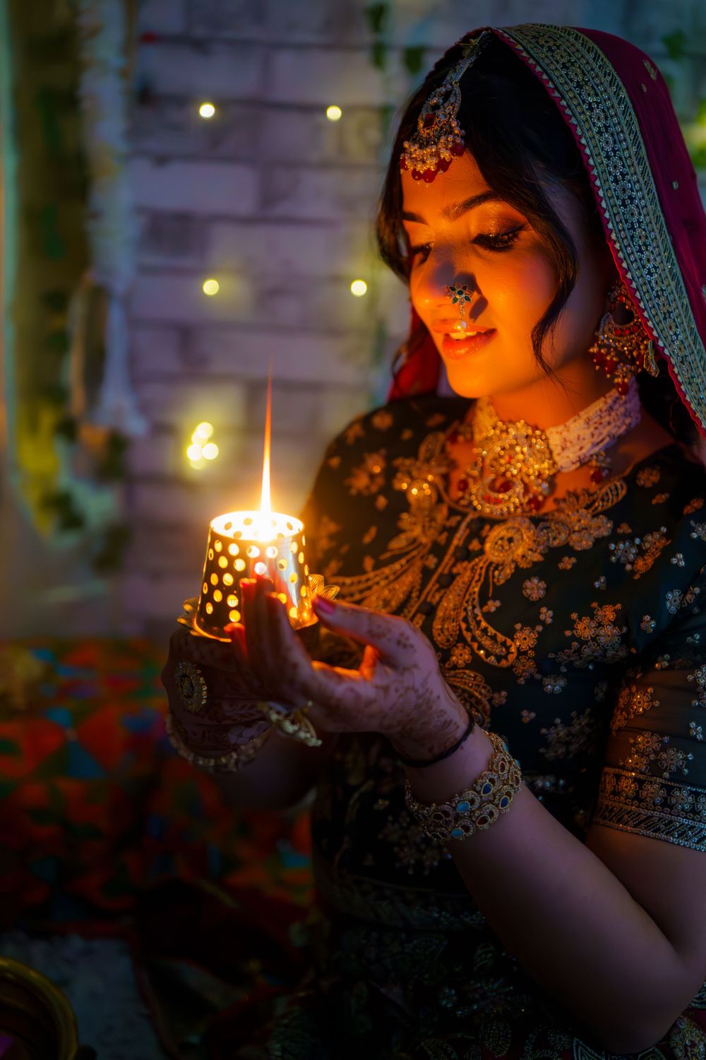Traditional bride in ornate embroidered dress and jewelry holds glowing candle creating warm atmospheric lighting.