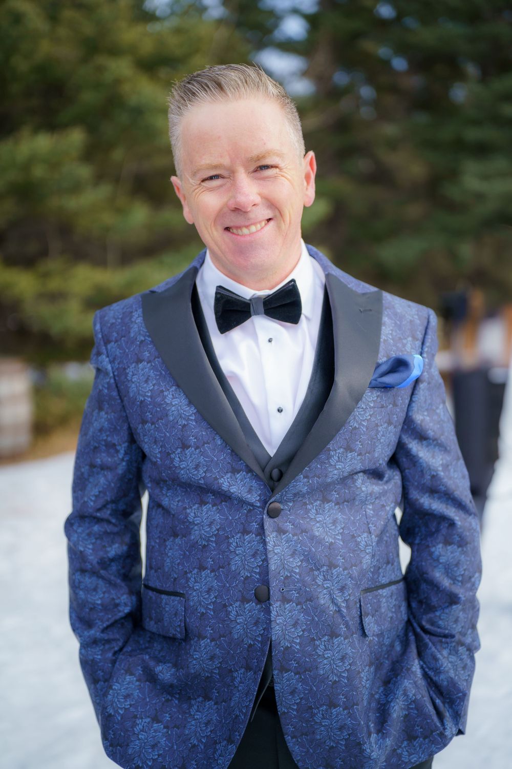 Someone wearing a formal blue patterned tuxedo jacket with black lapels and bow tie stands outside in snowy winter setting.