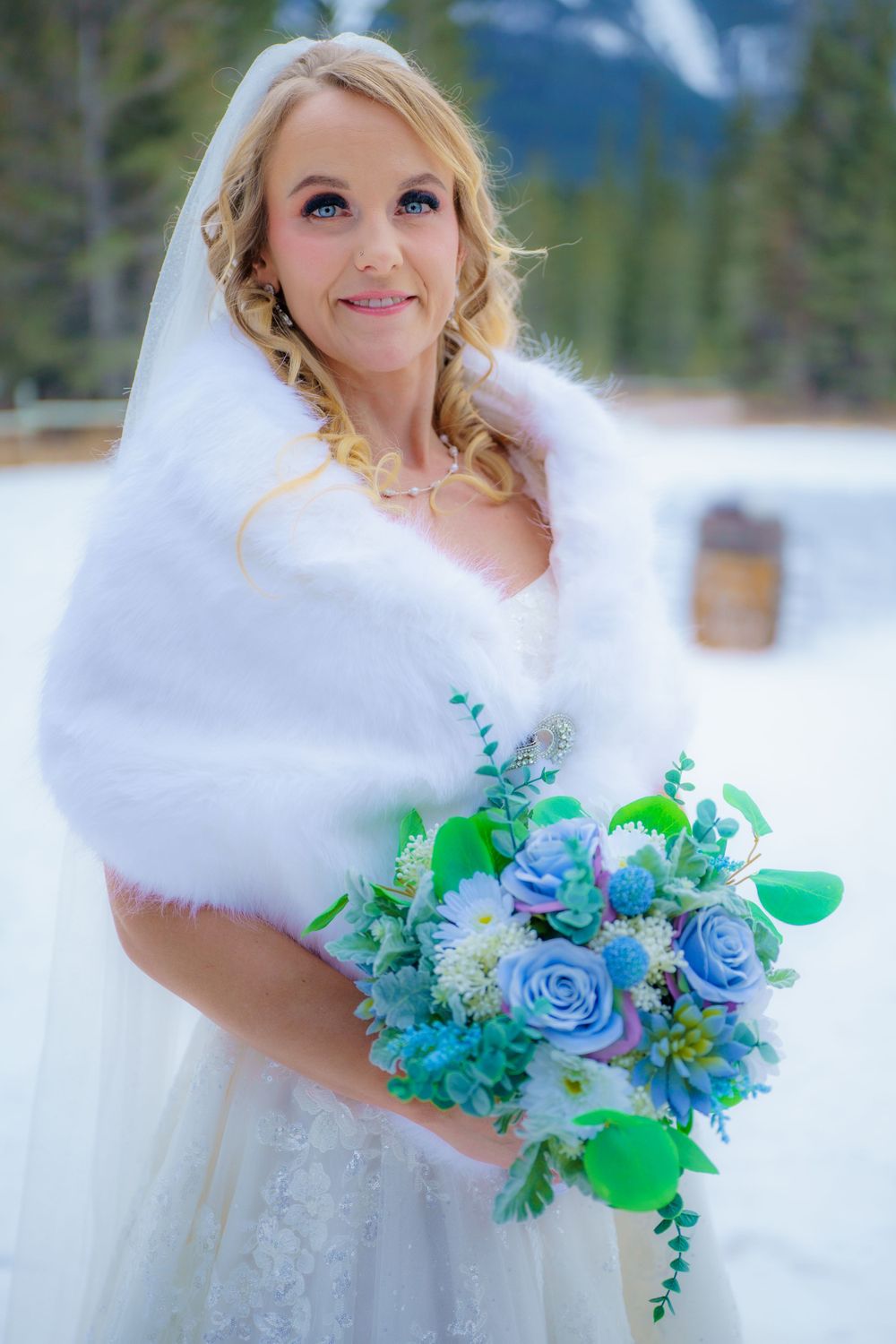 A bride in a white dress and fur shawl holds a vibrant blue and turquoise floral bouquet against a snowy mountain backdrop.