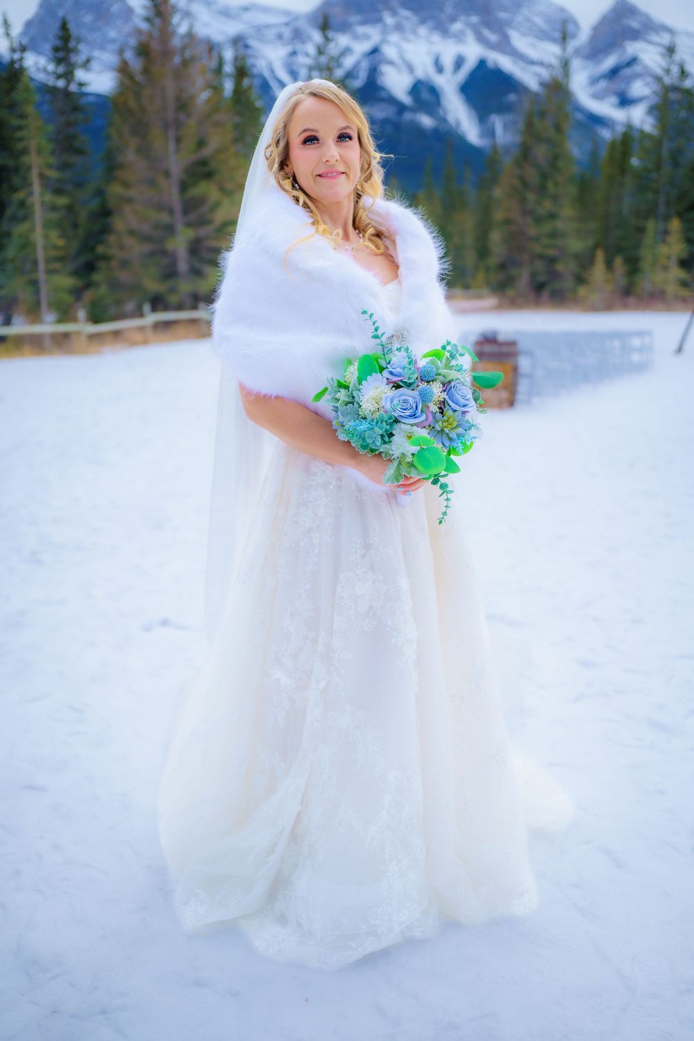 A winter bride in a white gown and fur shawl holds blue flowers against a snowy mountain backdrop in Canadian Rockies.