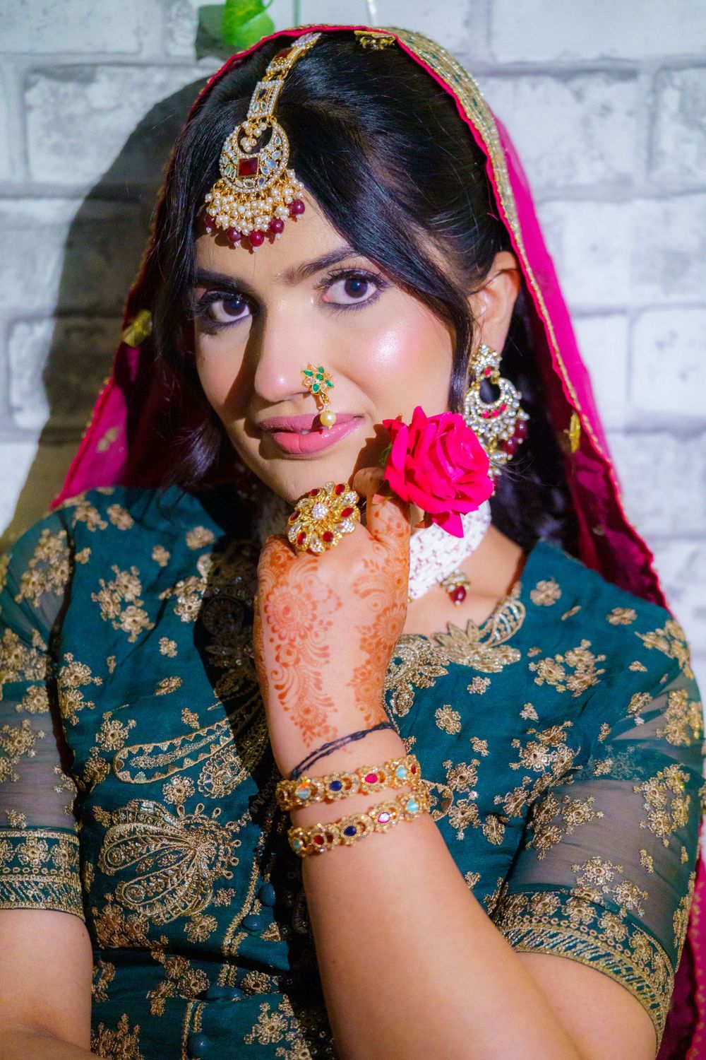 Traditional Indian bridal attire with teal and gold embroidered dress, pink veil, and ornate jewelry showcased against brick wall.