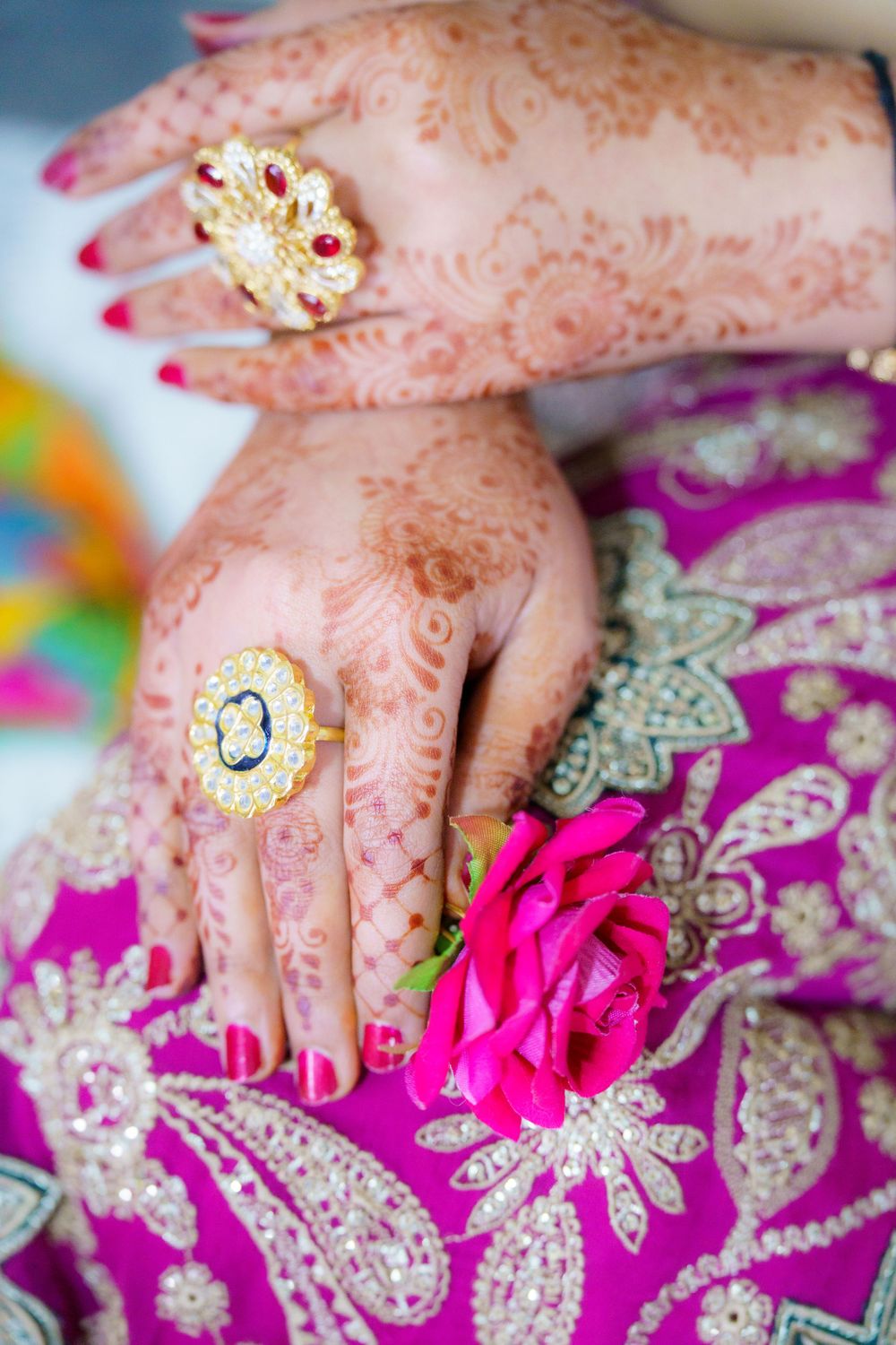 Hands adorned with intricate henna designs and gold rings rest on vibrant pink floral fabric with a magenta rose.