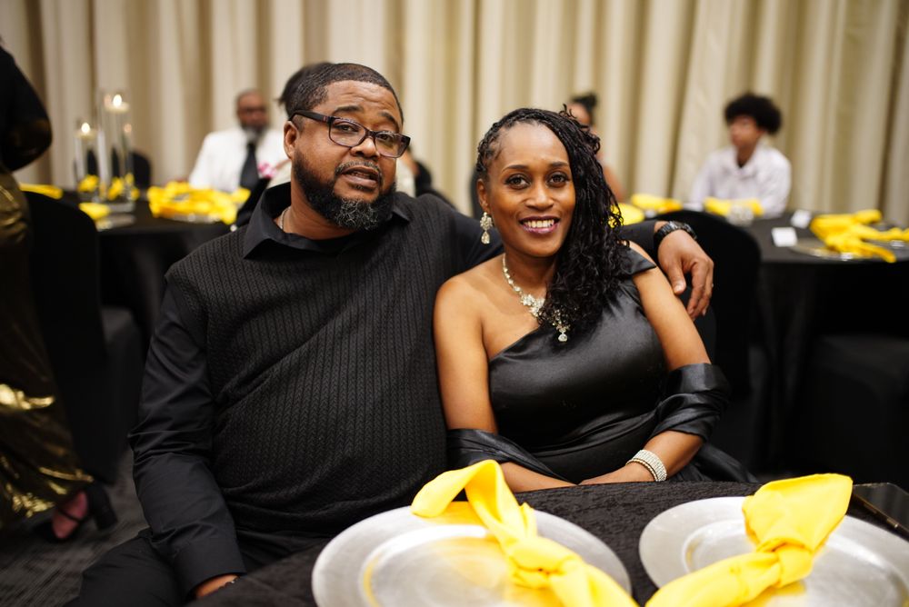 Two people smile while sitting at a table decorated with yellow accents during an indoor event.
