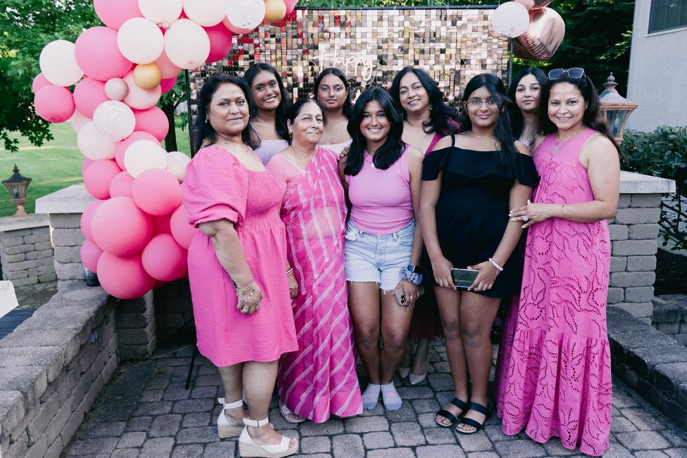 A group in coordinated pink outfits poses together by stone steps with pink balloon arch decorations.