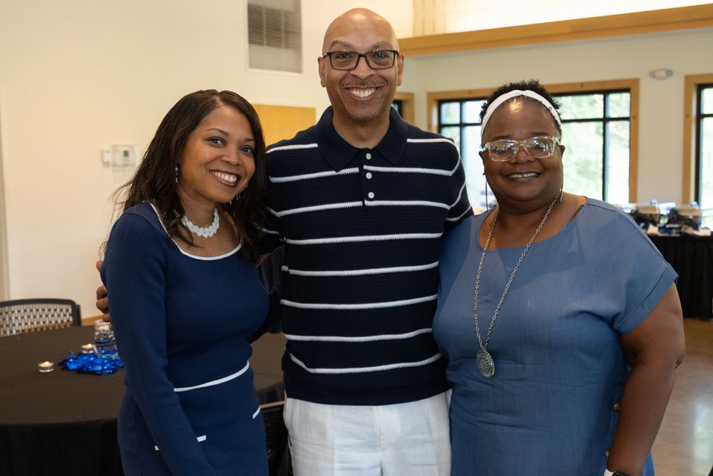 Three people stand together smiling in an office or community space wearing business casual attire.
