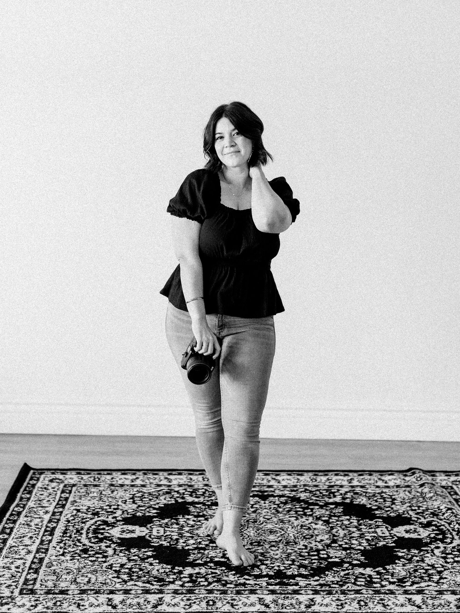 A black and white photo shows someone in a black top and jeans standing barefoot on a patterned rug.