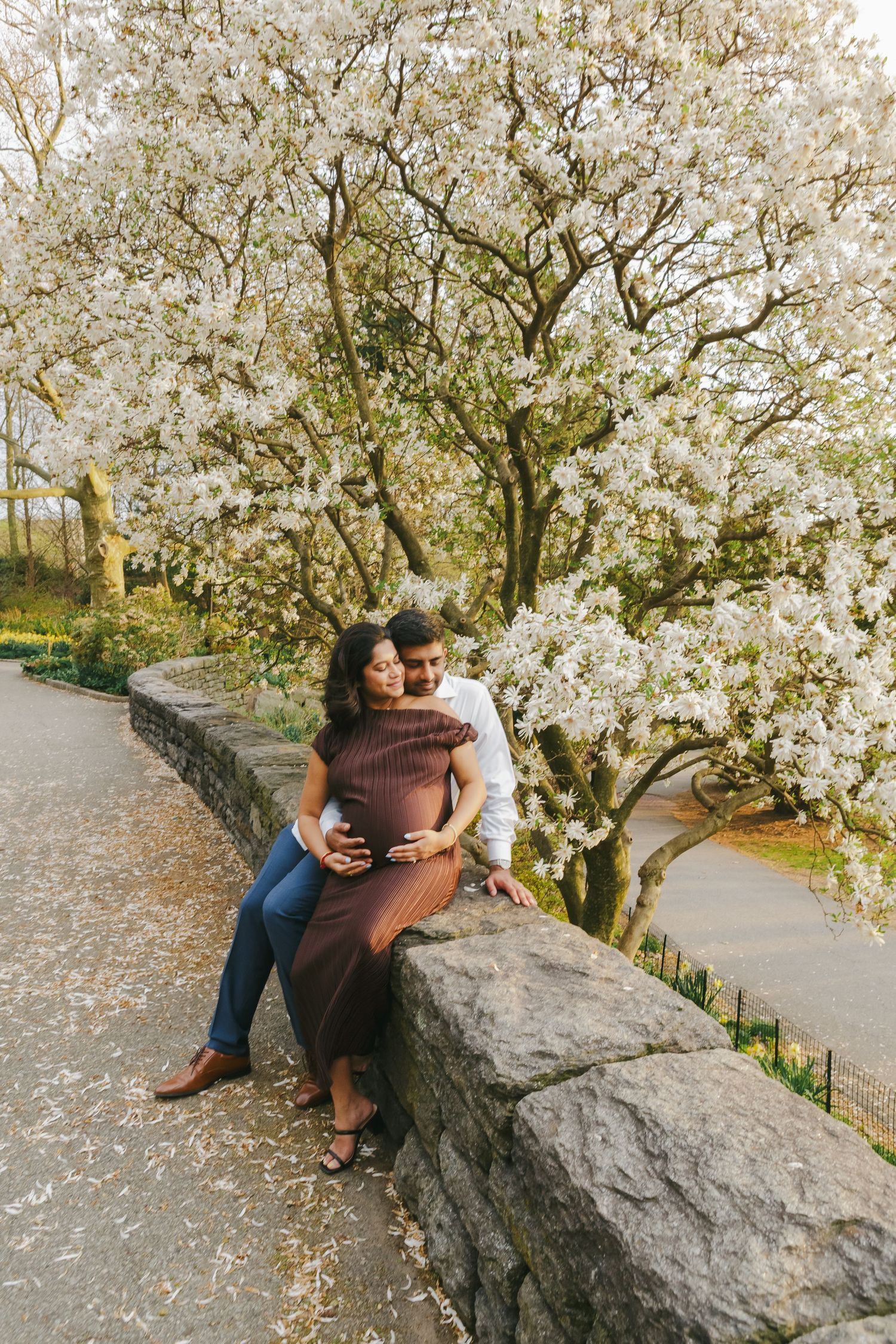 Expecting parents illuminated by golden light, holding hands under cherry blossoms, captured at sunset.