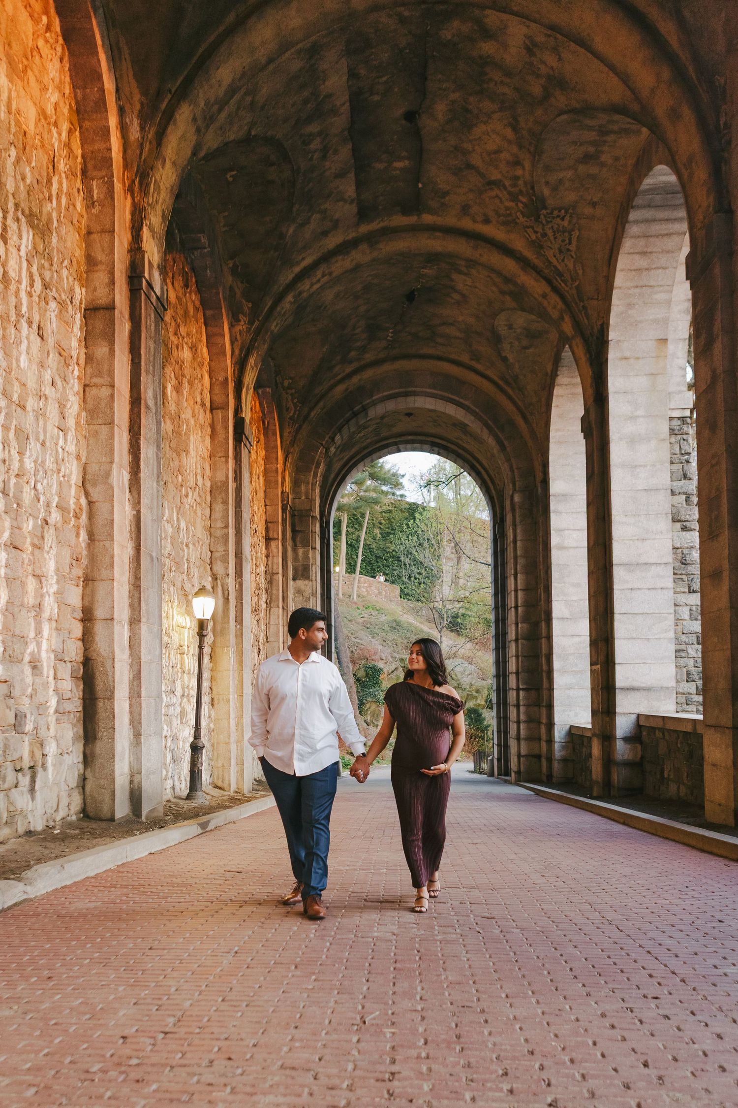 Expecting couple standing beneath Fort Tryon Park’s grand arches, embraced by soft golden-hour light in an elegant maternity portrait.