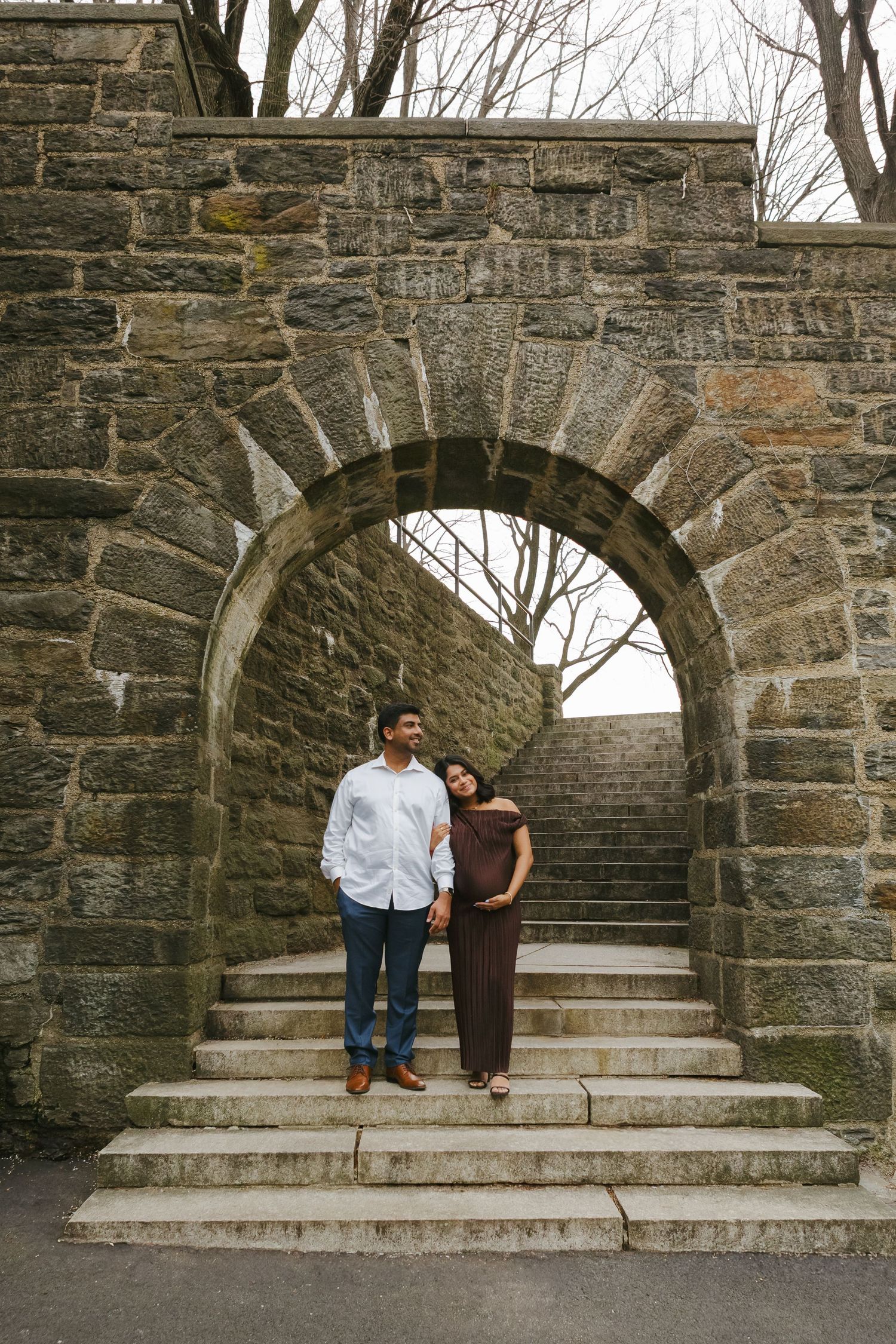 Expecting couple gazing at each other, surrounded by the golden glow of sunset at Fort Tryon Park, by NYC maternity photographer Paula B