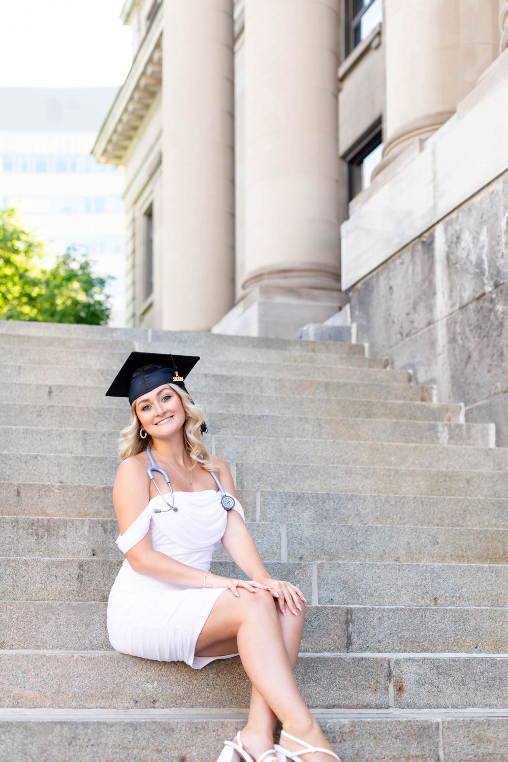 Graduate in white dress sitting on stone steps of classical building.