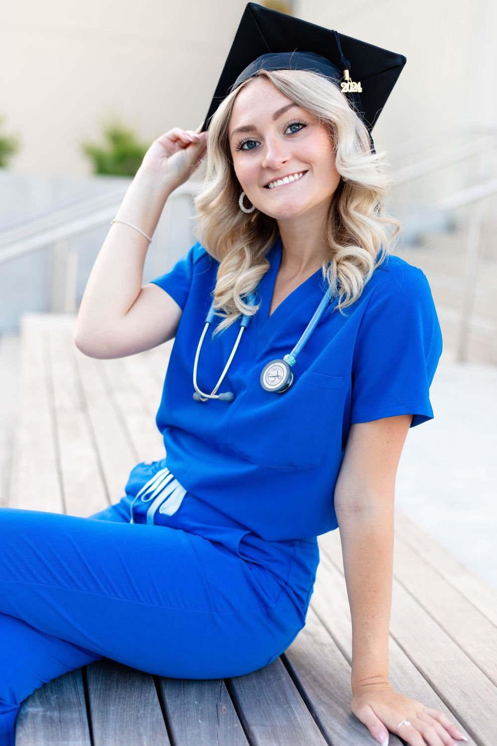 Graduate in blue medical scrubs wearing cap and stethoscope smiles while sitting outdoors.
