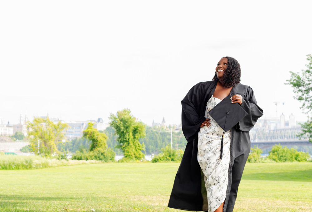 Woman in flowing dress and black coat poses outdoors in a park setting.