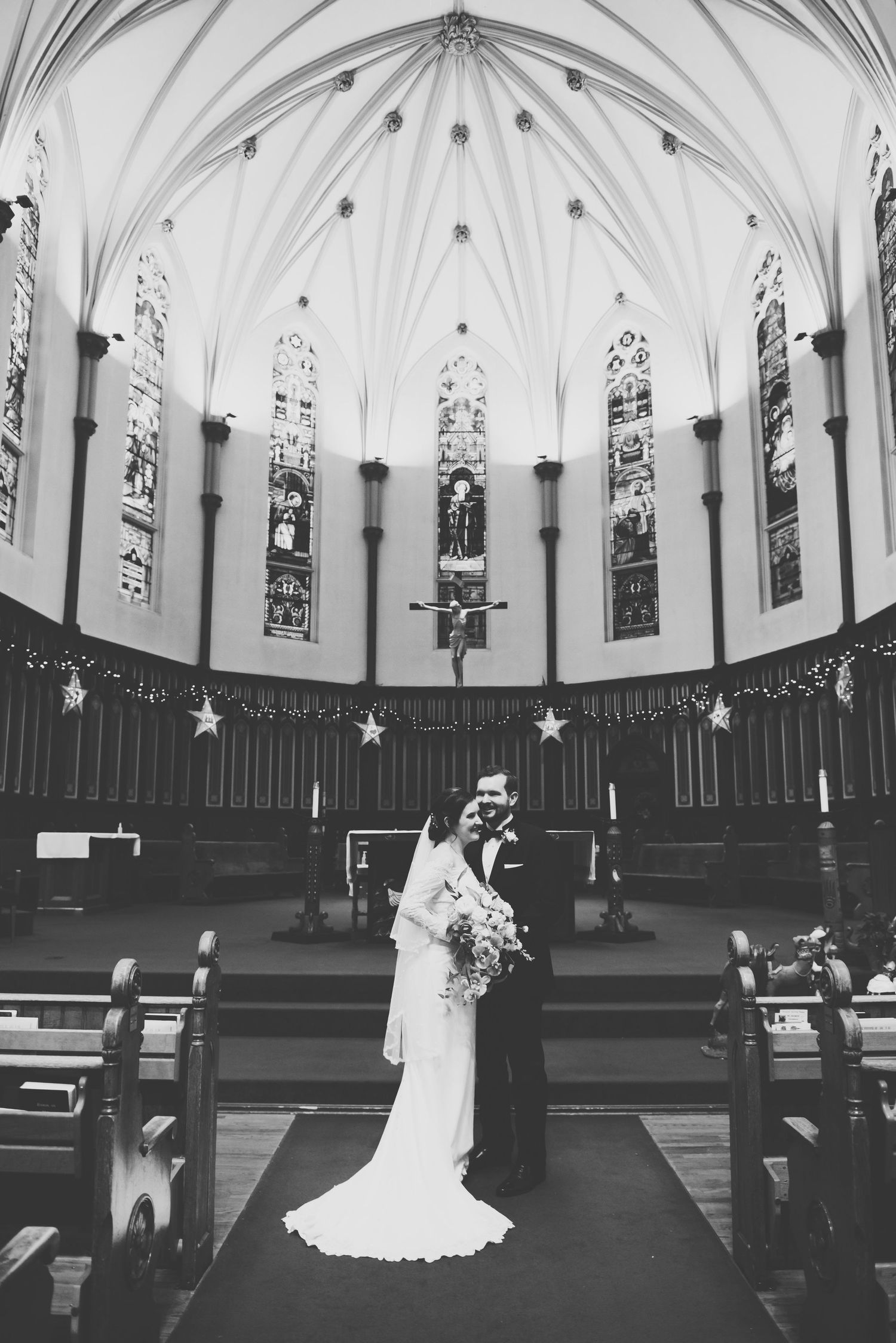 Black and white photograph of a couple on their wedding day embracing in a church with gothic arched windows and vaulted ceilings.