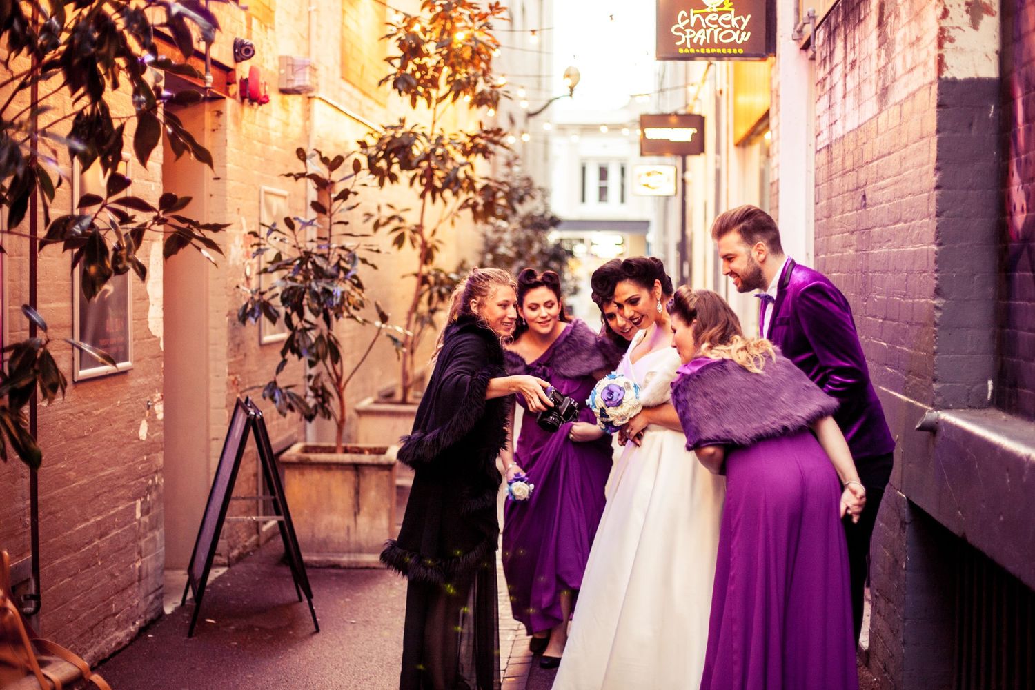 A candid wedding moment shared with guests wearing purple dresses in an urban alleyway.