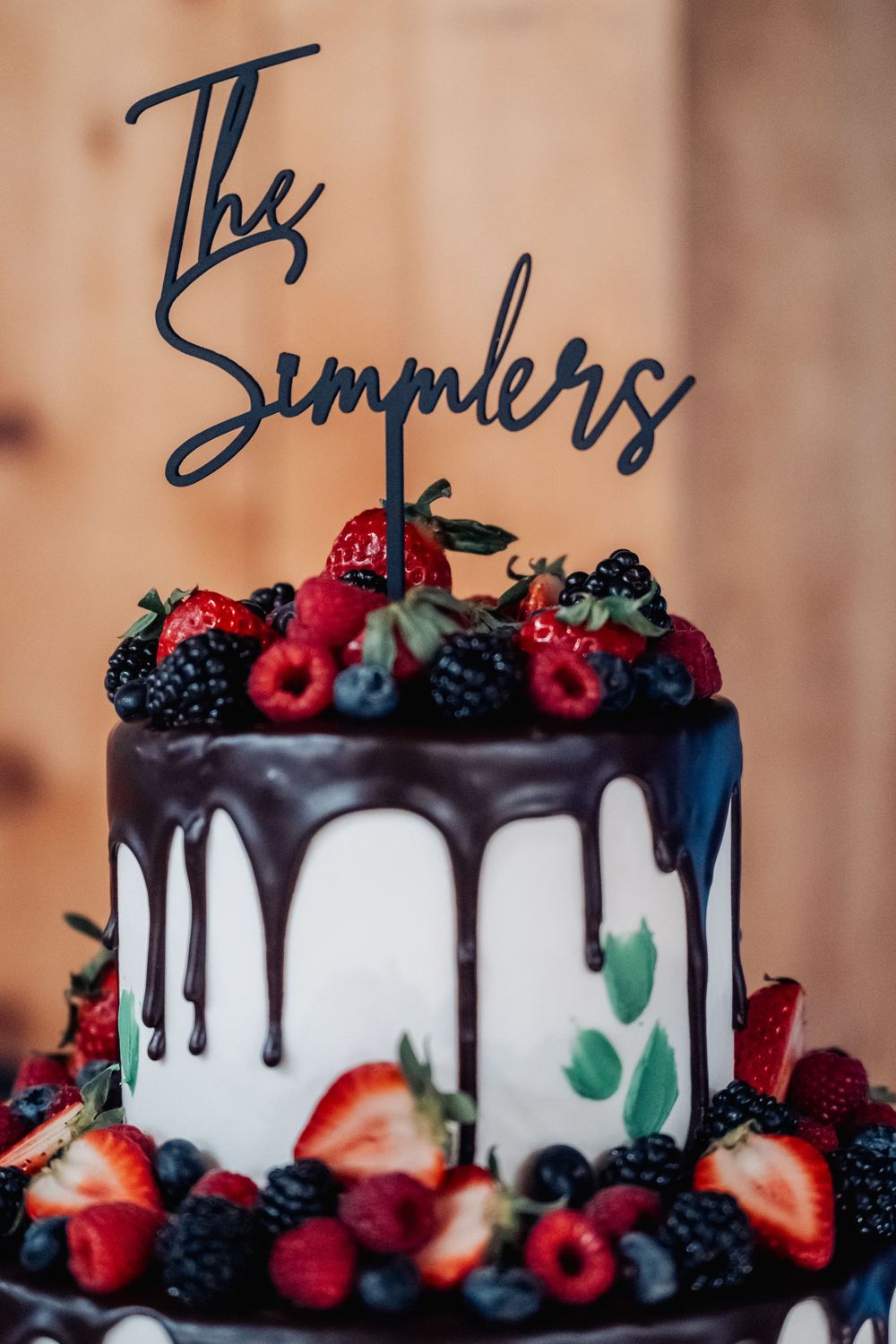 White wedding cake with chocolate drips topped with fresh berries and a custom black 'The Simpers' topper decoration.