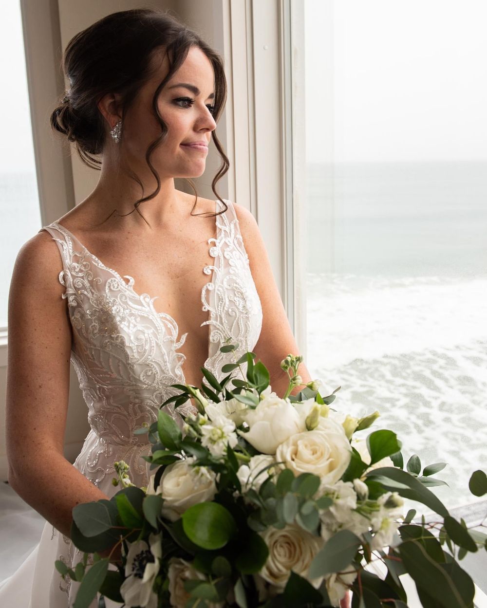 Bride holding white and green bouquet poses by window in lace wedding gown.