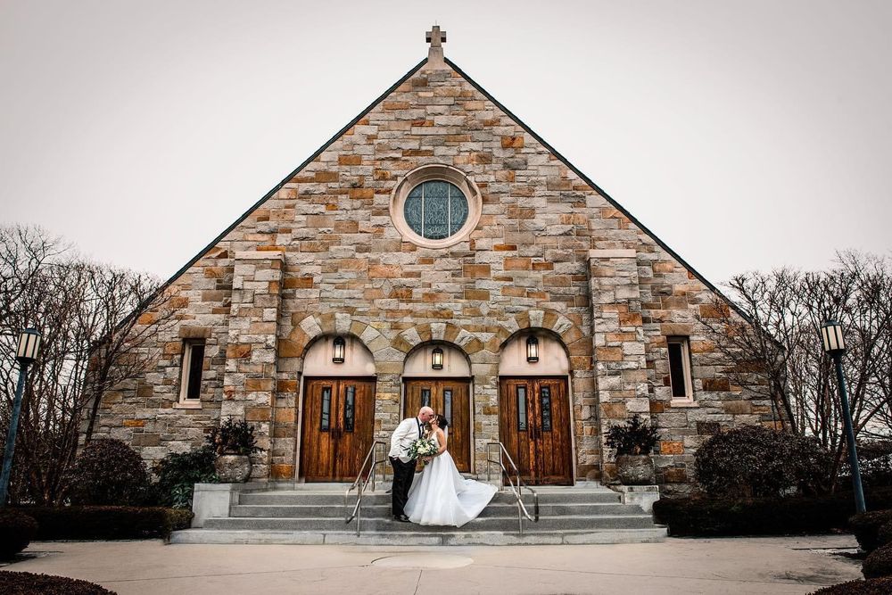 Wedding couple poses in front of stone church with arched wooden doors.