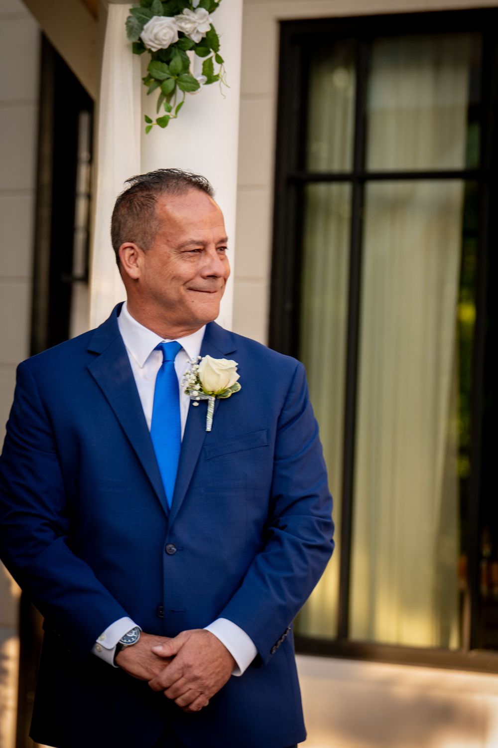 A groom in a navy blue suit with bright blue tie and white boutonniere smiles while standing outside a building at a wedding.