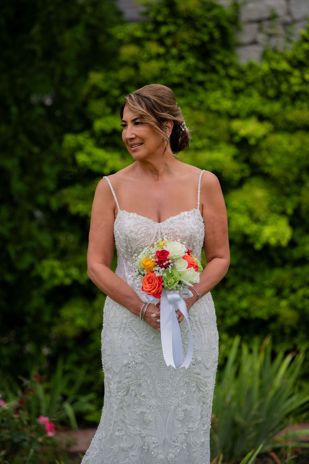 Bride in lace spaghetti strap wedding gown holds vibrant bouquet of coral and yellow flowers in garden setting.