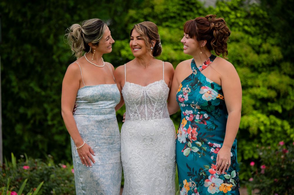 Three women wearing elegant formal dresses smile together in a garden setting with green foliage in the background.