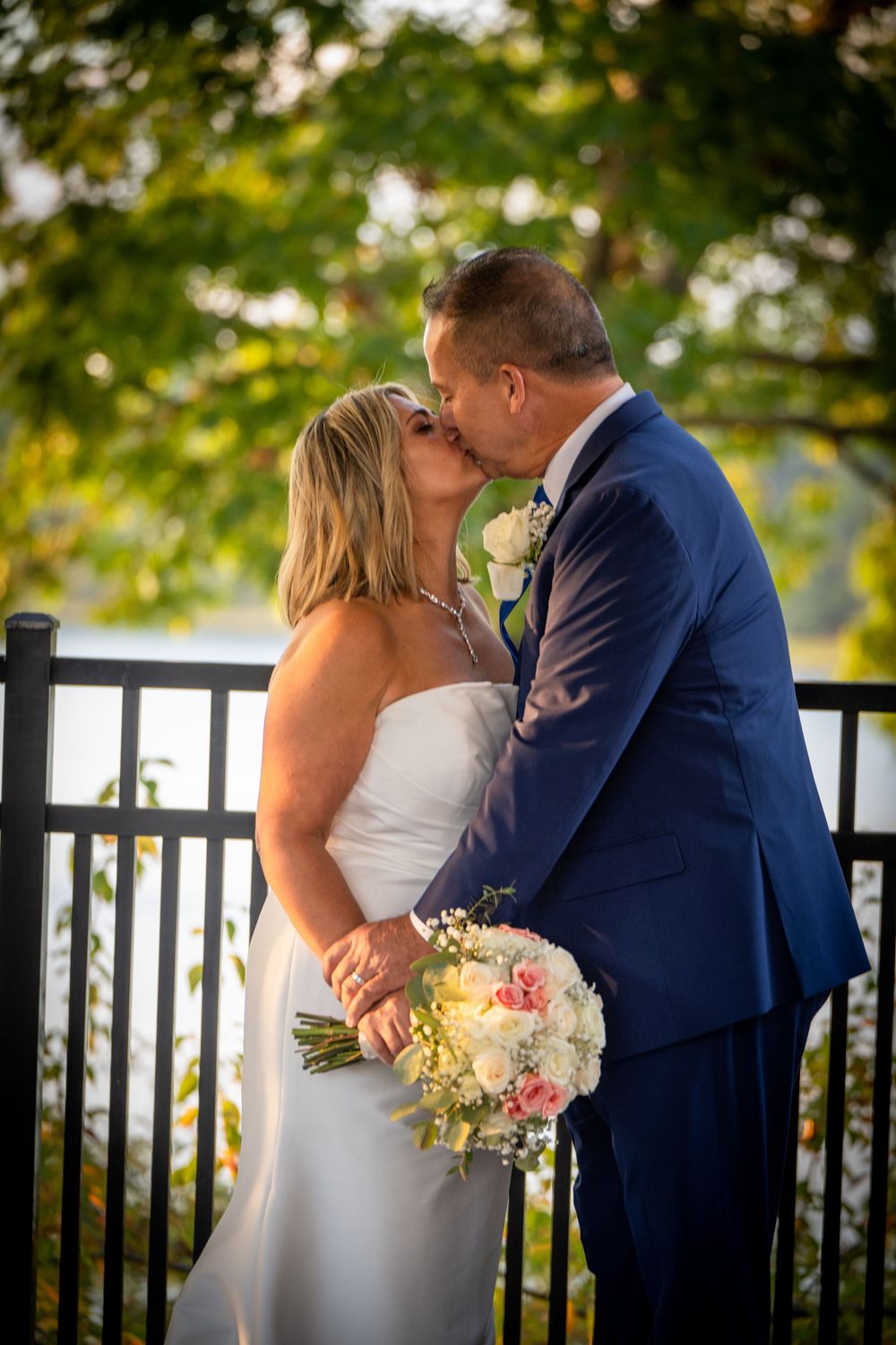 Romantic wedding couple shares a kiss on a deck railing while holding a pastel bouquet at sunset.