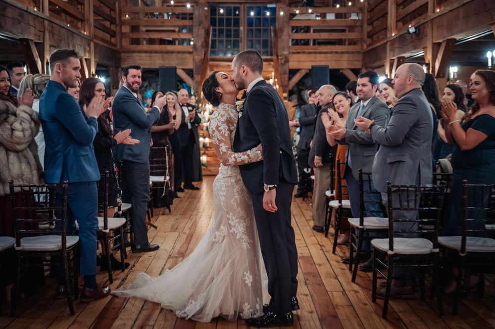 Wedding ceremony inside a rustic wooden barn venue with guests applauding the newlyweds during processional.
