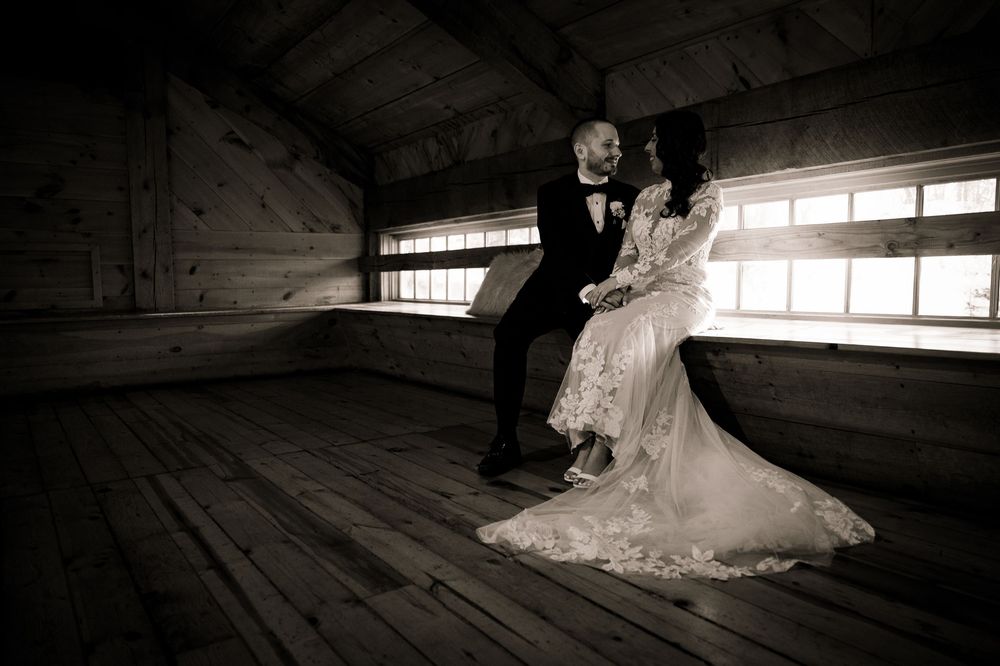 A bride and groom share a joyful moment while sitting together in a rustic barn with window light streaming in.