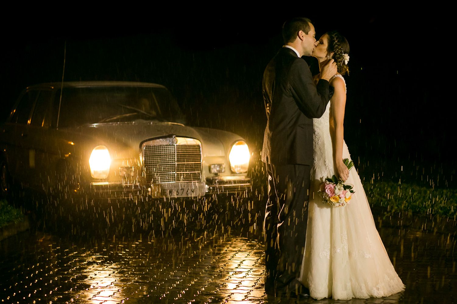 A couple embraces in front of a vintage car with glowing headlights on a rainy evening.
