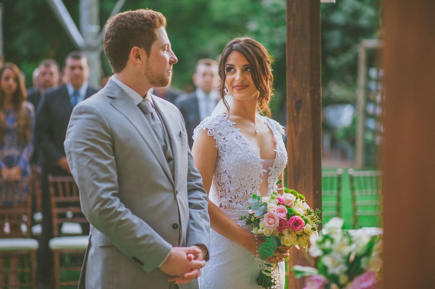 A bride in a white lace gown holds a pink bouquet while standing with a groom in a gray suit at an outdoor ceremony.
