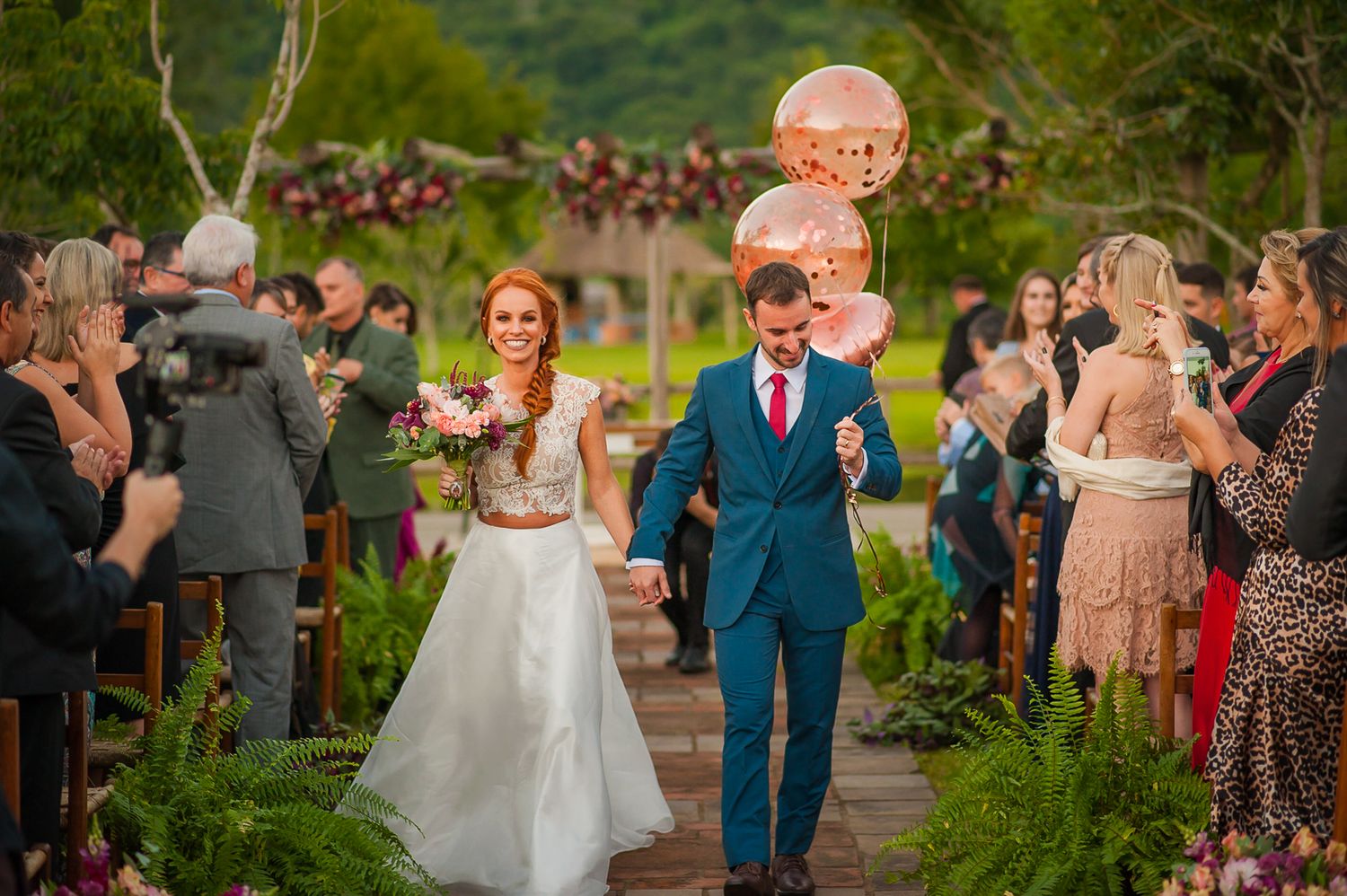 A wedding celebration outdoors with a happy couple walking down a garden path as guests look on during sunset.